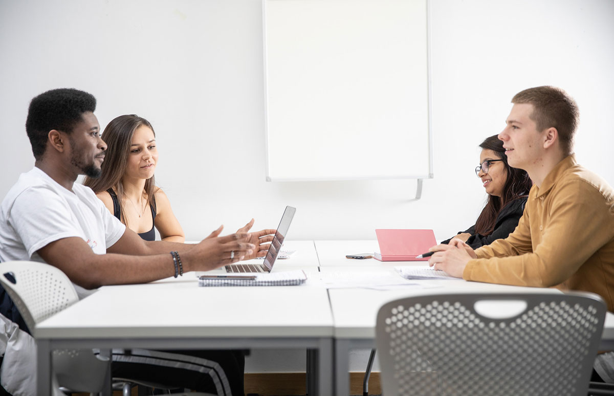four students sit around a table in a white classroom. an open laptop and notebooks can be seen on the table, the students are talking