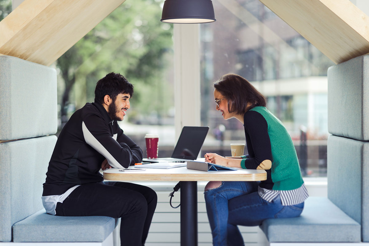 Two students sit in a booth in the university library across the table from each other. a coffee cup, open laptop and notepads can be seen on the table. the students are smiling