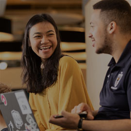 Male and female students chatting to each other and laughing. The female student is wearing a yellow jumper and the male student is wearing a navy hoodie.