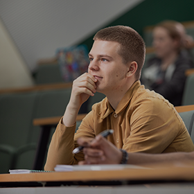 A student with short hair and a yellow shirt is sat in a lecture theatre