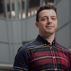 Close-up of a male student wearing a checkered button shirt, stood in the middle of the Business School