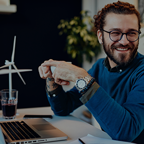A guy is sat at his desk holding a cup of tea and laughing. He is wearing a blue jumper and has his laptop open.