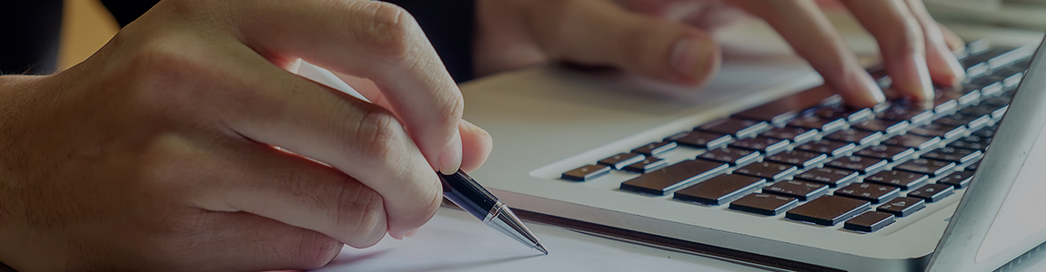 Close-up of an individual making notes on a pad with a ballpoint pen and using a laptop with his other hand