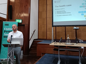 Professor Matthew Johnson pictured giving a keynote address, standing in front of a lecturn with a large screen beghind him