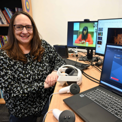 Professor Nic Whitton pictured sitting at a desk next to several computer screens and VR headsets