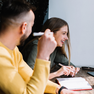 A man in a yellow jumper laughing with a women in a green jacket. She is writing in a note book.