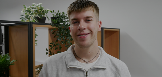 Student in grey hoodie, stood in front of plants, smiling