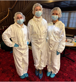 Three people wearing white protective suits, hairnets, face masks, gloves, and blue shoe covers standing on a red carpeted floor indoors.