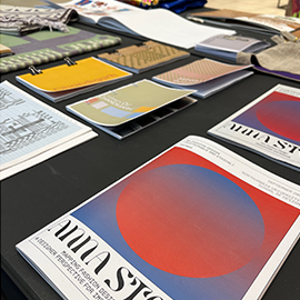 A table display featuring various design portfolios and booklets. The materials include colourful booklets with graphic covers, textile samples, and printed design sheets showcasing different aspects of design work.